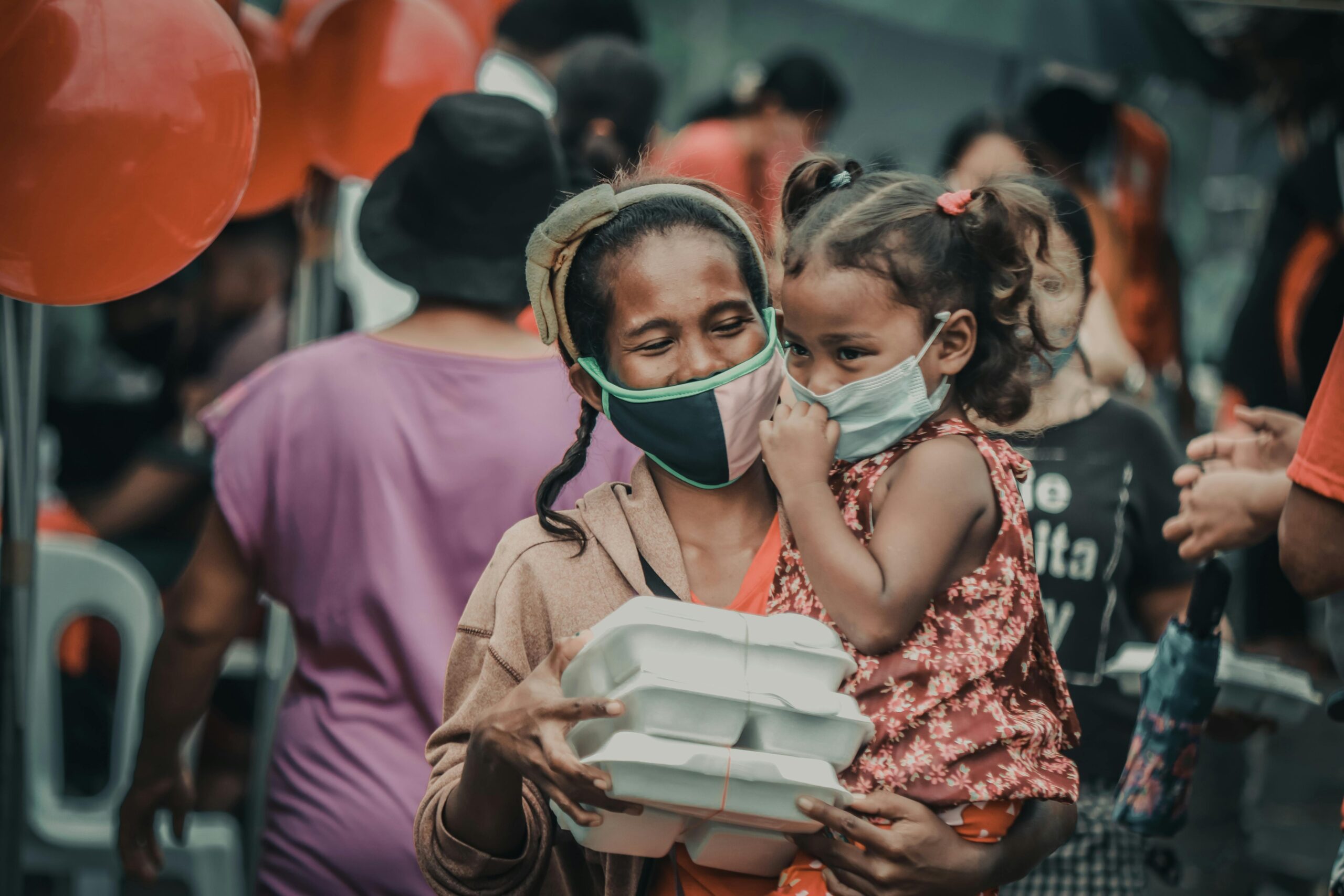 A mother and child wearing masks receive food packages at an outdoor event with balloons.